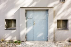 A heavy, light-blue $\text{guardian steel door}$ with a concrete frame flanked by two small, grated windows on a light gray stucco building.
