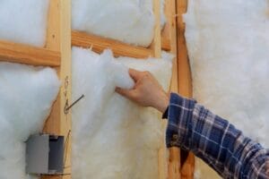 A close-up shot of a hand in a blue plaid shirt installing white fiberglass batt insulation between wood studs, a process similar to the steps detailing how to insulate a garage door for winter.