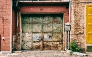 A very old, rusty, and heavily damaged garage door on a brick building, showing signs of severe wear.