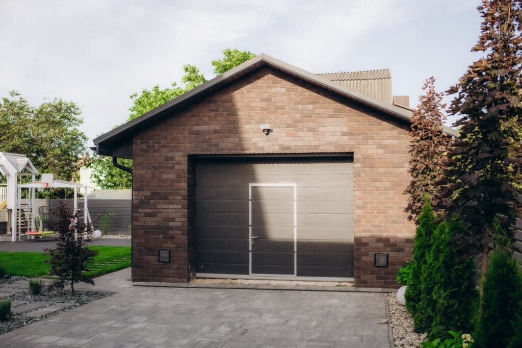 A modern, insulated dark gray garage door with a pedestrian door insert on a brick home with landscaping.