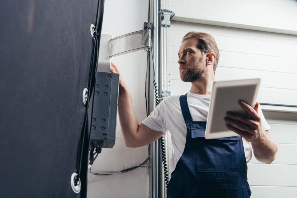 A homeowner holds a tablet while inspecting the electrical components of a garage door system.