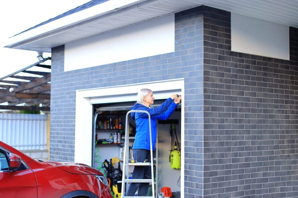 A woman on a stepladder performing a DIY garage door repair, tightening a bolt with a screwdriver.