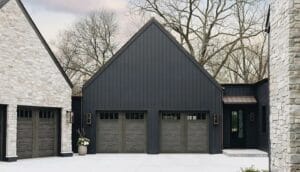 A large modern house featuring dark gray board-and-batten siding and two matching wood-look garage doors, suggesting a cold weather setting where a garage door frozen shut might be an issue.