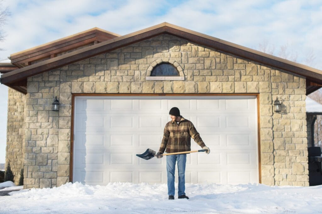 A man holding a snow shovel stands in front of a wide white garage door set in a stone-faced garage on a snowy day, preparing to clear the area where a garage door frozen shut might be stuck.