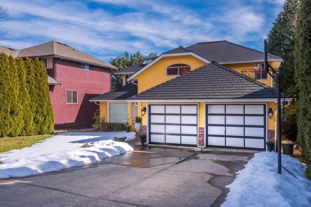 A bright yellow house with two modern white and black panel garage doors, framed by snow on the driveway and lawn, illustrating a common winter scenario where a garage door frozen shut can occur.