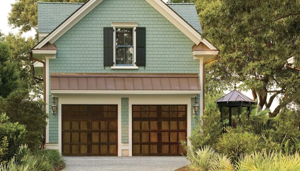 Shingled coastal-style home with light teal siding, copper awning, and two dark faux wood garage doors with square window panels, enhancing curb appeal.