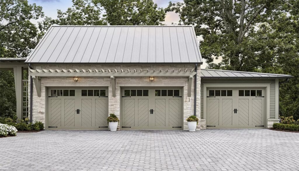 Modern farmhouse garage with three light sage green carriage house doors featuring chevron panels, black hardware, a light gray metal roof, and stone facade.