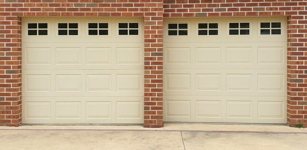 Two matching cream-colored insulated garage doors with small square windows are set in a red brick house.