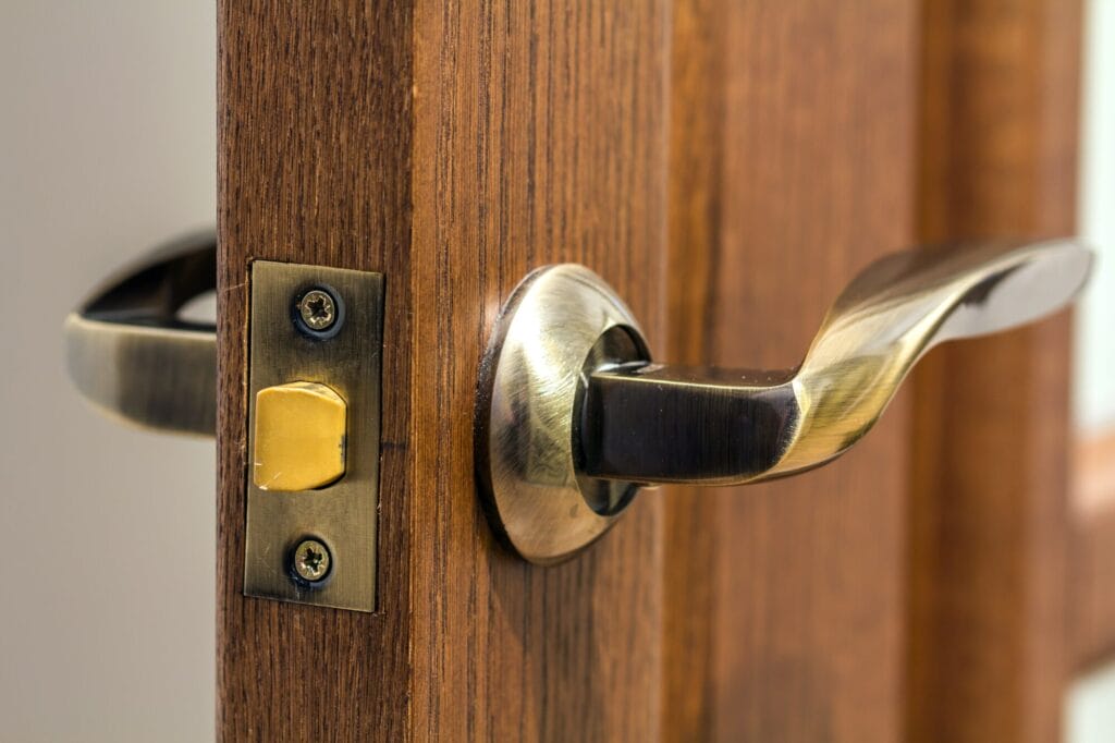 A close-up of an antique brass-colored door handle and its latch mechanism on a wooden door, showing some wear around the screws.