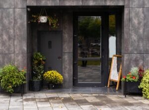 A modern building entrance with a dark gray door and a full-length glass side panel, accented by potted plants.