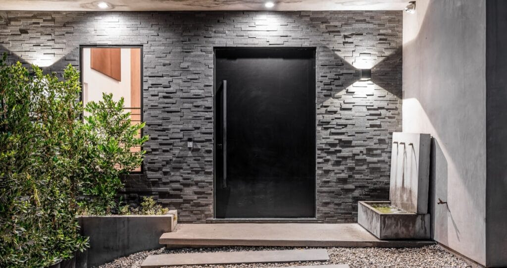 A modern, minimalist black steel front door set against a dark gray stacked stone wall, with a small window and concrete water feature.