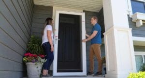 A man and a woman are standing on a front porch next to a newly installed white storm door with a black insert. The man is holding the door open while the woman looks on. The house has gray siding and white trim.