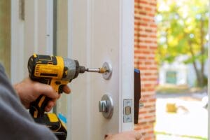 Close-up of a worker using a yellow and black cordless drill to remove a screw from a deadbolt mechanism on a white door during a deadbolt lock repair service.