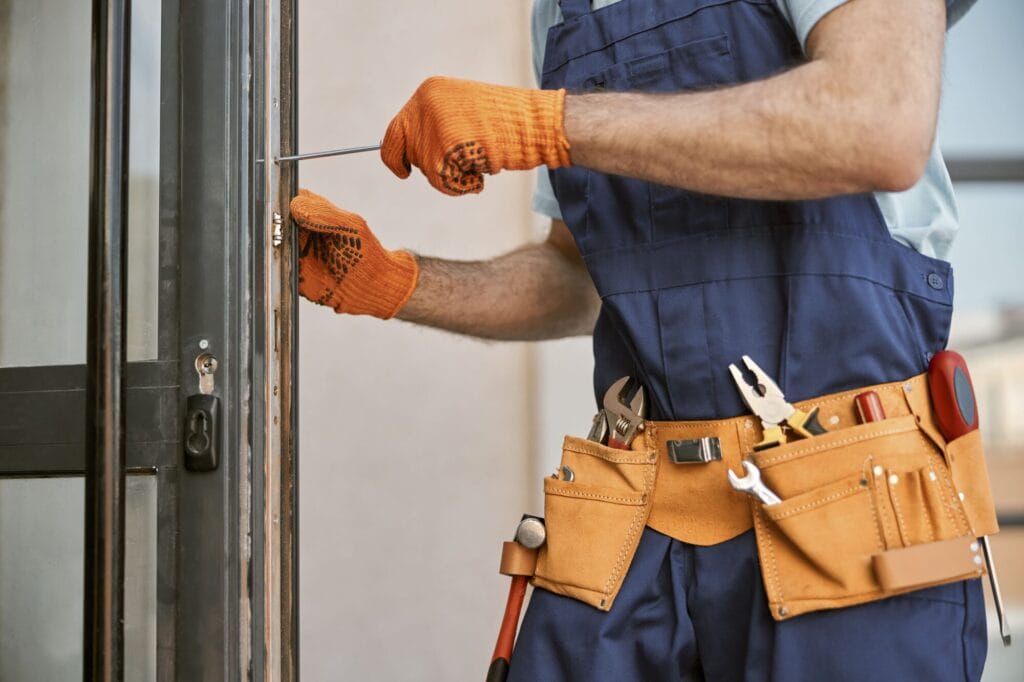 A close-up of a worker in blue overalls, an orange tool belt, and orange gloves using a screwdriver to adjust a lock mechanism on a metal doorframe, underscoring the hands-on expertise of an emergency locksmith.