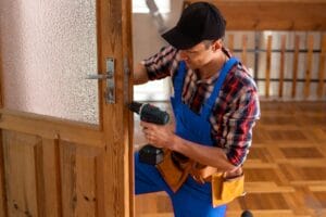 A man wearing blue overalls and a plaid shirt, identified as an emergency locksmith, uses a cordless drill to repair or replace a lock mechanism on a wooden door with a textured glass panel.