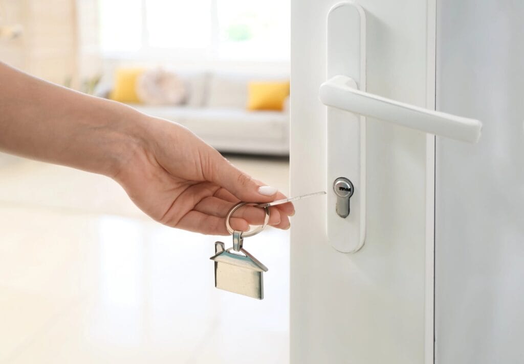 A person's hand inserts a house key, attached to a house-shaped keychain, into the cylinder of a modern white door handle, illustrating the variety of types of door locks used in residential settings.