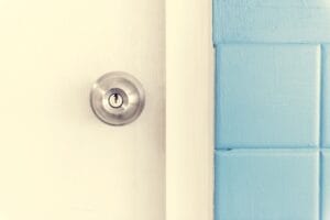 A close-up of a brushed metal keyed doorknob installed on a white door next to a blue tiled wall.