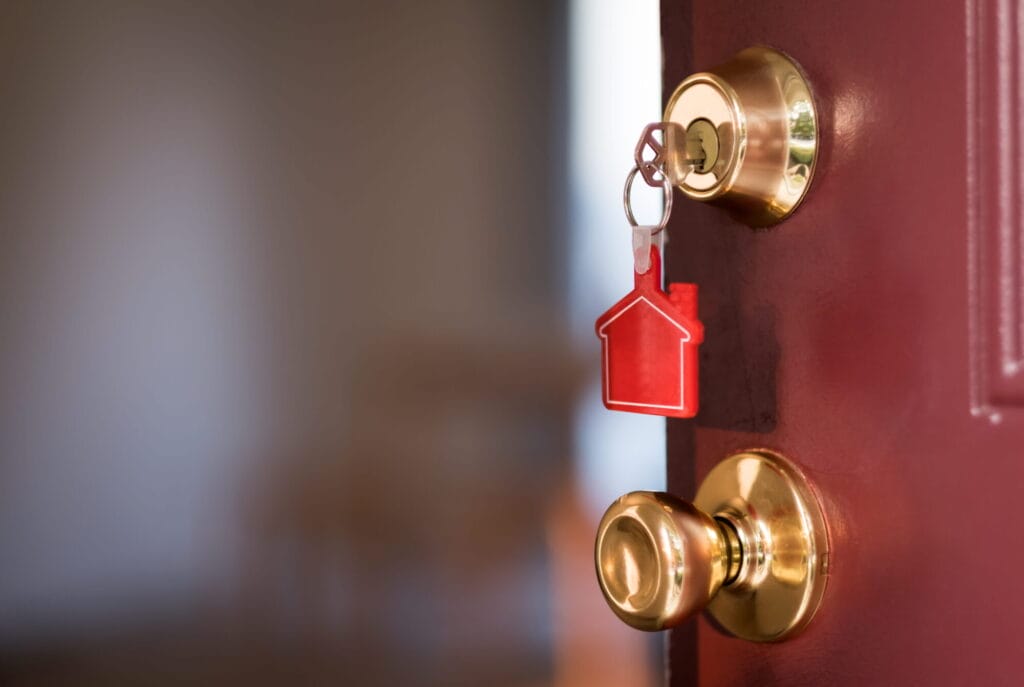 Alt Text: A close-up of an open burgundy door featuring a traditional polished brass deadbolt and matching doorknob, with a physical key and a red house-shaped keychain inserted into the lock.