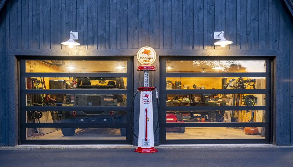 A dark blue exterior features a vintage gas pump standing between two large glass garage doors that display a brightly lit workshop and parked vehicle inside.