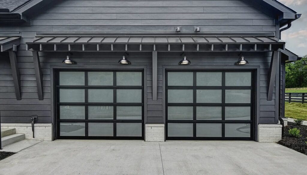 Two modern glass garage doors with black frames and frosted glass panels are set into a dark grey siding wall beneath a metal awning with four gooseneck lights.