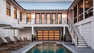 A sleek white farmhouse-style home featuring a poolside patio and a black-framed modern glass garage door that opens to a glowing interior space.
