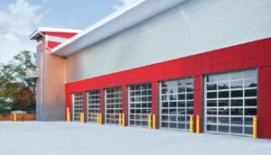 A row of modern commercial glass garage doors with silver aluminum frames installed on a red and grey industrial building.