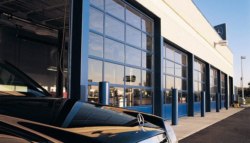 A luxury car dealership service bay featuring a series of blue-framed commercial glass garage doors with clear tempered glass panels.