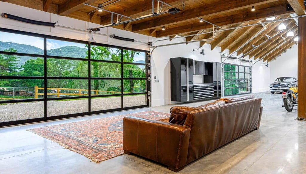 The interior view of a luxury garage featuring a brown leather sofa and large, clear panel full view glass garage doors that offer an unobstructed view of a scenic mountain landscape.