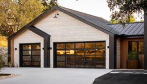 A modern home featuring sleek black full view glass garage doors installed against a white stone facade and dark metal roofing.