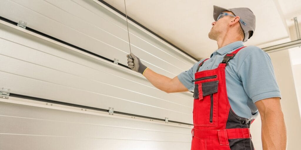 A technician in red overalls and safety glasses looks up while pulling a release cord along the garage door track.