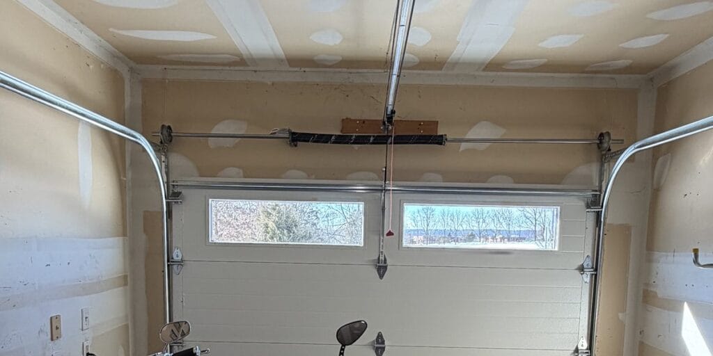 Looking up inside a garage, metal tracks run along both walls and curve overhead into a horizontal rail above a light colored door with two window panes, motorcycles parked underneath