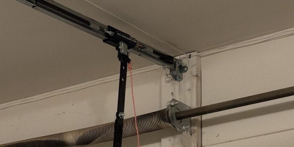 A metal spring bar and support bracket bolted into a wood header board above a garage door, with a red pull cord hanging down from an angled black arm