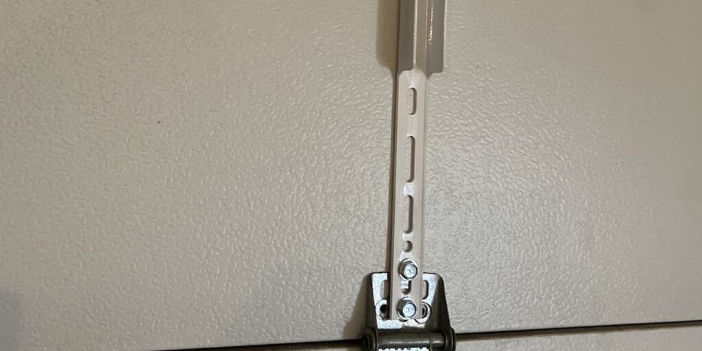 A slotted metal reinforcement bracket runs down a garage wall and bolts into a hinge plate near the bottom of the door track.